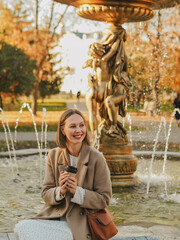 Beautiful blonde young woman in hat and coat drinking coffee and walking in autumn city