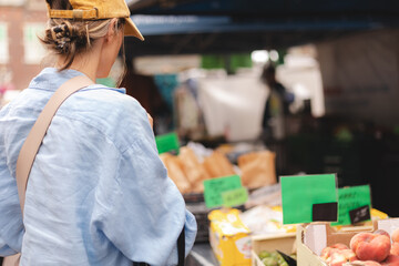 Female customer dressed casual standing at greengrocery's market and choosing fresh organic vegetables fruits. Back of a young woman purchasing fresh raw products and groceries at framer's market.