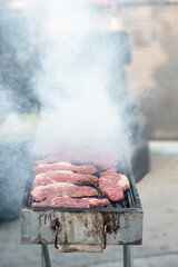 Steaks are being grilled on a metal barbecue, surrounded by billowing smoke. The activity takes place in a backyard setting, perfect for a casual gathering.