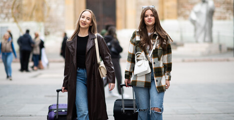 Two stylish young women walking outdoors with suitcases, exploring an urban destination during...