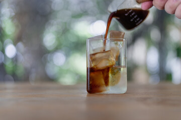 Hand pouring coffee into a glass tumbler with coffee with ice, on a wooden table with a nature background, the shot is taken from a frontal angle