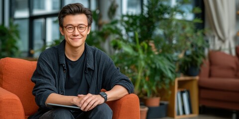 Obraz premium Young man with glasses smiling, seated on an orange sofa in a cozy, plant-filled modern living space.