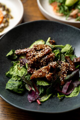 A vibrant salad sits enticingly on a dark plate, featuring fresh greens topped with savory meat pieces drizzled in a rich sauce and garnished with sesame seeds