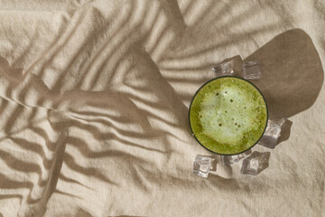 Summer day scene with iced matcha latte and tropical palm tree leaf shadow on sand color linen background. Sun and shadows. Minimal summer matcha latte concept. Flat lay style. Top view.