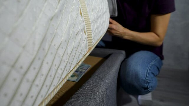 Close-up of a hand taking a stack of US dollar bills under a mattress bed. Hiding or saving money