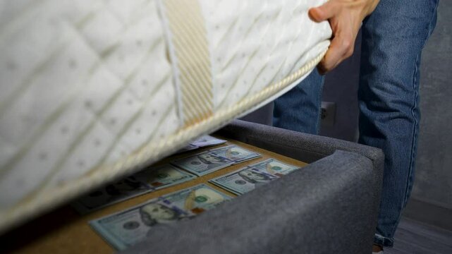 Close-up of a person hiding a stack of US hundred-dollar bills under a mattress