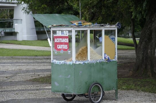 An old green popcorn cart with a red sign "we acept card" stands under a large tree in a park.
