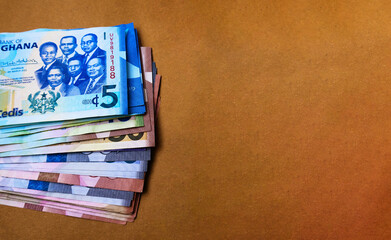 Various denominations of Ghanaian cedi currency notes stacked at the side of screen on brown...