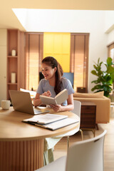Young Asian woman working with laptop and book at home office. Happy female learning online, studying or distance education, remote worker in apartment.