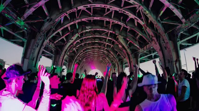 A DJ performs under a bridge at night, surrounded by a crowd. Low-angle shot captures the dynamic, energetic vibe, perfect for a music video.