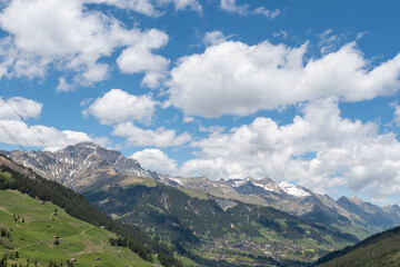 Soft spring clouds drift across a bright blue sky, casting gentle shadows over the alpine valley. Below, the village of Adelboden rests quietly, surrounded by awakening nature.