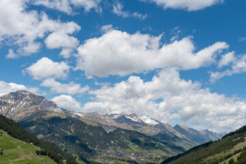 Soft spring clouds drift across a bright blue sky, casting gentle shadows over the alpine valley....