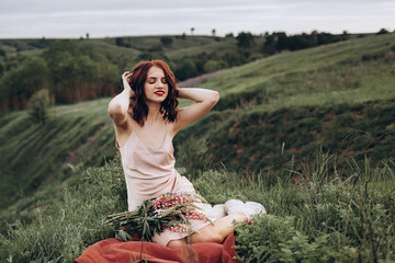 A beautiful red-haired young woman in a pink dress is sitting in nature among flowers at sunset. The girl is smiling. In the background, there are hills and ravines.