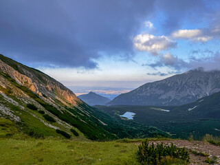 This breathtaking image captures a serene mountain valley landscape at twilight, showcasing lush hills, a reflective lake, and a dramatic cloud-filled sky