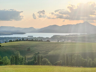 This stunning landscape features rolling hills, a serene lake, and majestic mountains, illuminated by the warm glow of the setting sun and soft rays of light breaking through clouds