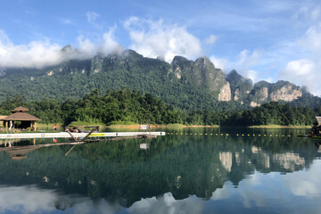 Stunning reflection of lush green mountains and a floating resort on a calm lake in Thailand. Morning mist hovers above the forested cliffs under a bright sky, creating a tranquil tropical escape.