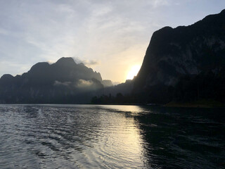 Golden sunlight breaks through misty limestone mountains at dawn, casting reflections over calm lake waters. A serene and moody landscape captures the beauty of Thailand's tranquil natural scenes.