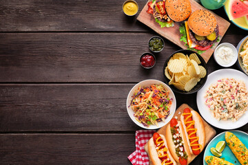 Summer food table scene with hot dogs, hamburger and sides. Top view side border over a dark wood  background. Copy space.