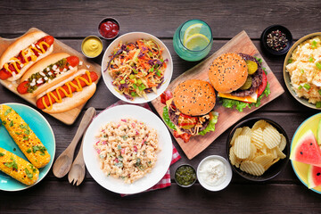 Summer food table scene with hot dogs, hamburger and sides. Top view over a dark wood background.