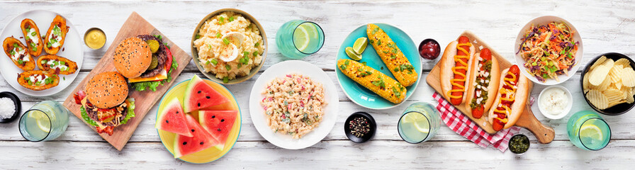 Summer food table scene with hot dogs, hamburger and sides. Above view over a white wood banner background. Copy space.