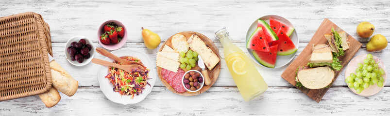 Summer picnic food table scene. Top view over a white wood banner background. Picnic basket, fruit, salad, sandwiches and snacks.