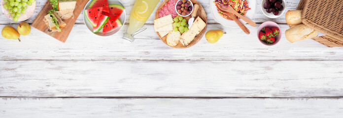 Summer picnic food top border. Top view over a white wood banner background. Picnic basket, fruit, salad, sandwiches and snacks. Copy space.