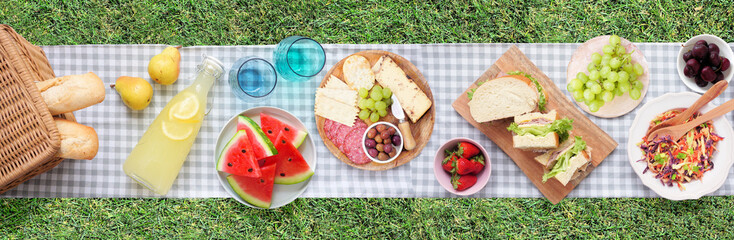 Summer picnic food scene. Top view with blanket over a green lawn banner background. Picnic basket, fruit, salad, sandwiches and snacks.