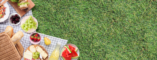 Summer picnic food corner border. Top view with blanket over a green lawn banner background. Picnic basket, fruit, salad, sandwiches and snacks. Copy space.