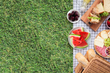 Summer picnic food side border. Top view with blanket over a green lawn background. Picnic basket, fruit, salad, sandwiches and snacks. Copy space.