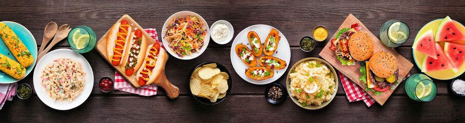 Summer food table scene with hot dogs, hamburger and sides. Top down view over a dark wood banner background.