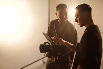Side angle of young videographer and assistant director analyzing footage on set during video...