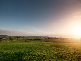 A large field of grass with a few houses in the distance. The sky is cloudy and the sun is setting. Calm and relaxing nature scene with rich color and warm summer vibe. Nobody. Picturesque view.