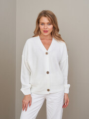 Woman wearing white knitted cardigan with button detail, standing confidently in front of a neutral...