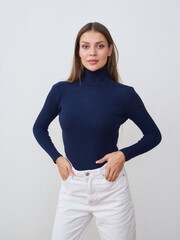 Woman with long hair wears a navy turtleneck and white pants, standing with hands on hips, gazing...