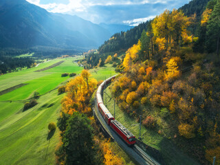 Aerial view of red train is moving in the swiss alps valley at sunset in autumn. Top view of...