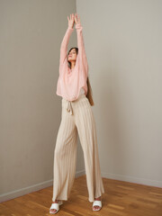Fototapeta premium Woman in pink shirt and beige wide-leg pants, stretching arms upward indoors on wooden floor. Her long hair flows down her back, creating a relaxed, elegant pose without shoes