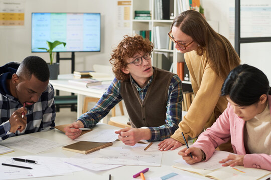Group of diverse students engaging in foreign language studies in classroom sitting around table filled with notes and books, with one supervisor assisting learners in improving their skills
