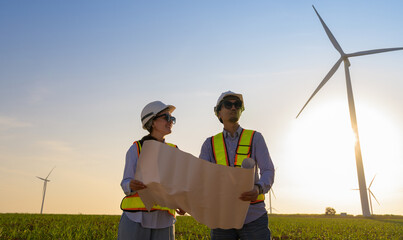 Wind farm engineers in high visibility vest and white helmet review blueprints for sustainable energy development at sunset while standing in green field with wind turbines