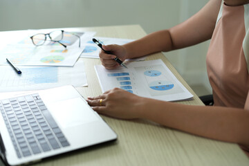 An asian young boss lady is busy working with laptop at home office and checking the paper documents of the company annual profits and sending email or SMS to her business partner using mobile phone.