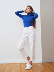 Woman with long hair in blue turtleneck and white pants standing against a light wall. She wears white sneakers and poses confidently with hands touching her head on hardwood floor
