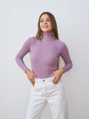 Woman smiling, wearing a lilac turtleneck sweater and white pants. Her hands are resting on her...