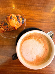 Close-up of a buttery croissant and a foamy cappuccino on a wooden table. Captured with a smartphone in a cozy Spanish café. A classic, comforting breakfast moment.