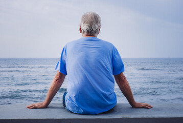 Rear view of senior retired man sitting at seaside looking at horizon over water enjoying freedom,...