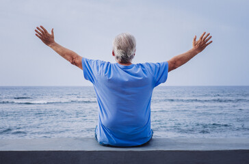 Rear view of senior retired man sitting at seaside looking at horizon over water enjoying freedom, vacation or retirement concept