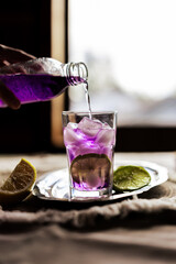 A woman pours a vibrant purple drink into a glass with ice and lime slices, evoking freshness and summer vibes. Ideal for beverage promotions, cocktail recipes