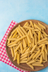 Uncooked penne pasta on a wooden plate with red checkered napkin, top view on blue background. Bright minimal food composition.