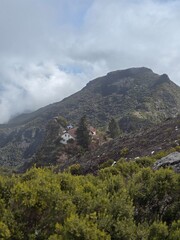 Fototapeta premium Clouds drift between the rugged peaks of Pico Ruivo, while a tiny house in the distance anchors the vast mountainous terrain.
