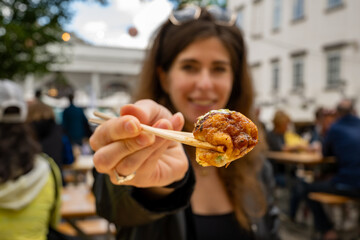 Young woman smiling while holding a takoyaki ball with chopsticks toward the camera. Close-up focus...