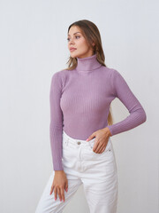 Woman in a light purple turtleneck sweater and white pants poses confidently, looking to the side....