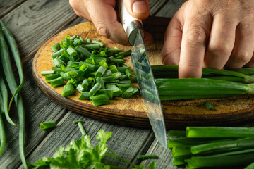 Hands skillfully slice green onions on a wooden cutting board surrounded by fresh herbs. The...